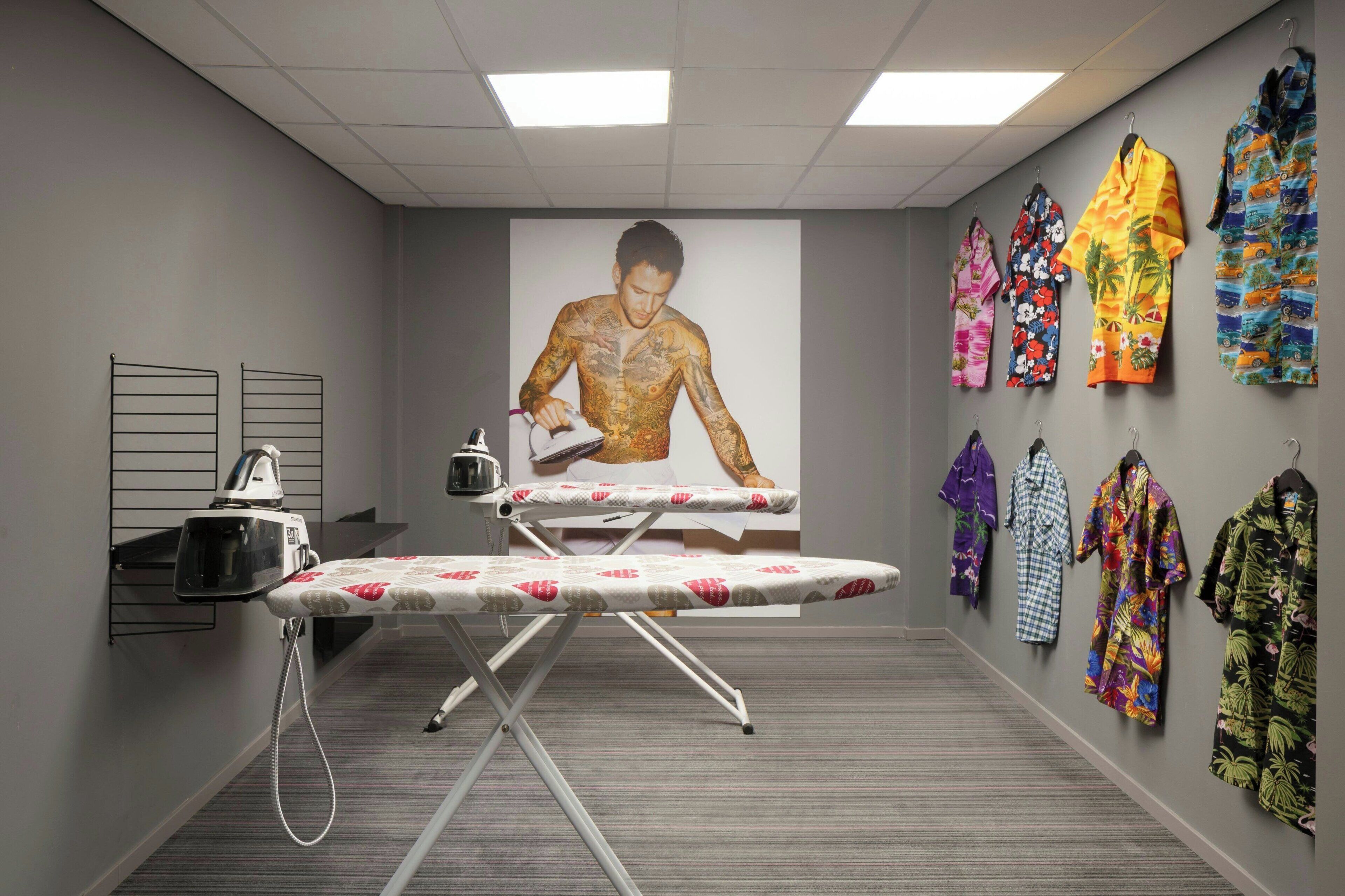 A room with an ironing station, a large poster of a shirtless man, and a wall displaying colorful robes.