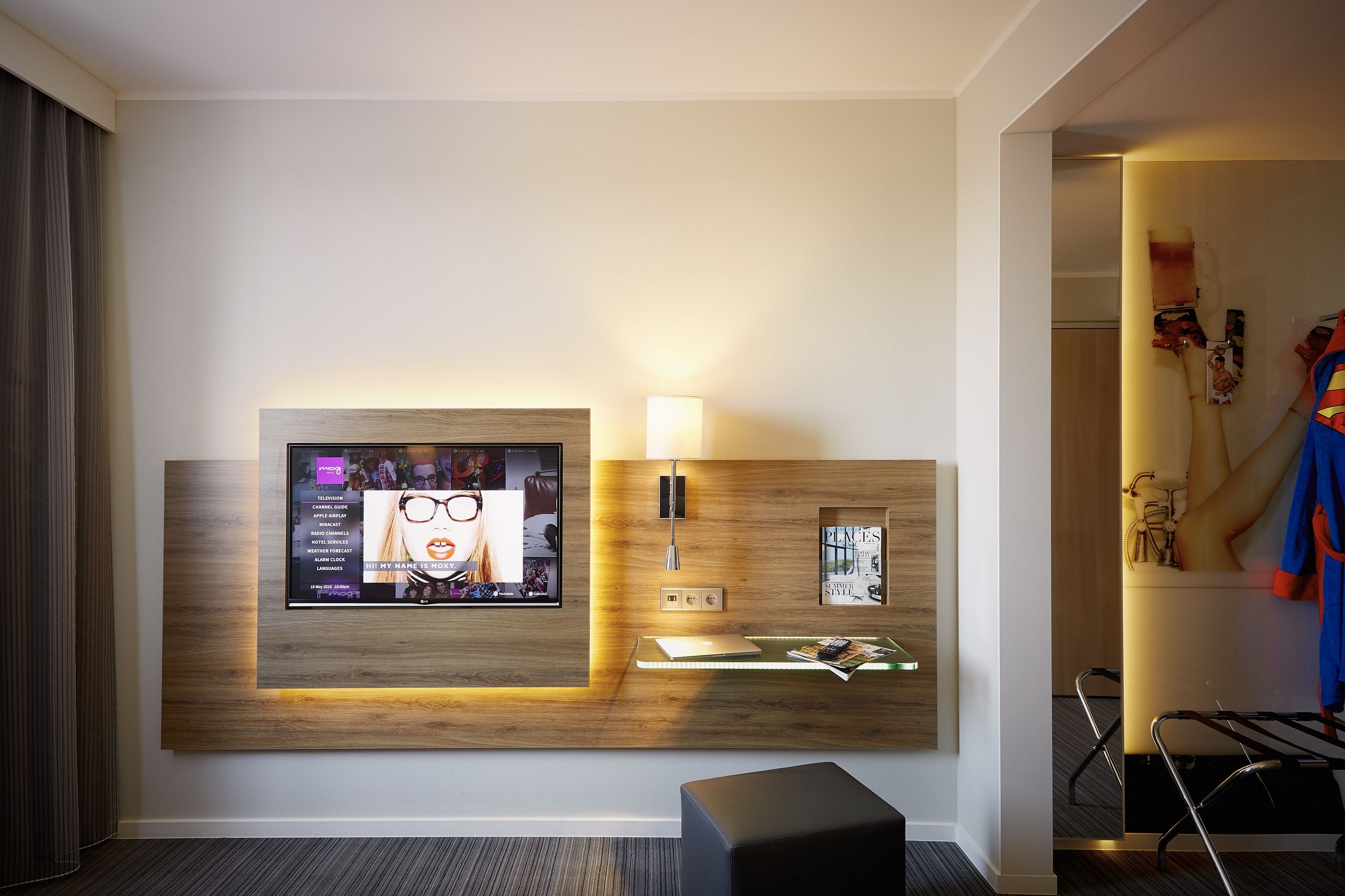 Habitación de hotel con televisor montado, lámpara de noche, revistero y perchero con una chaqueta.