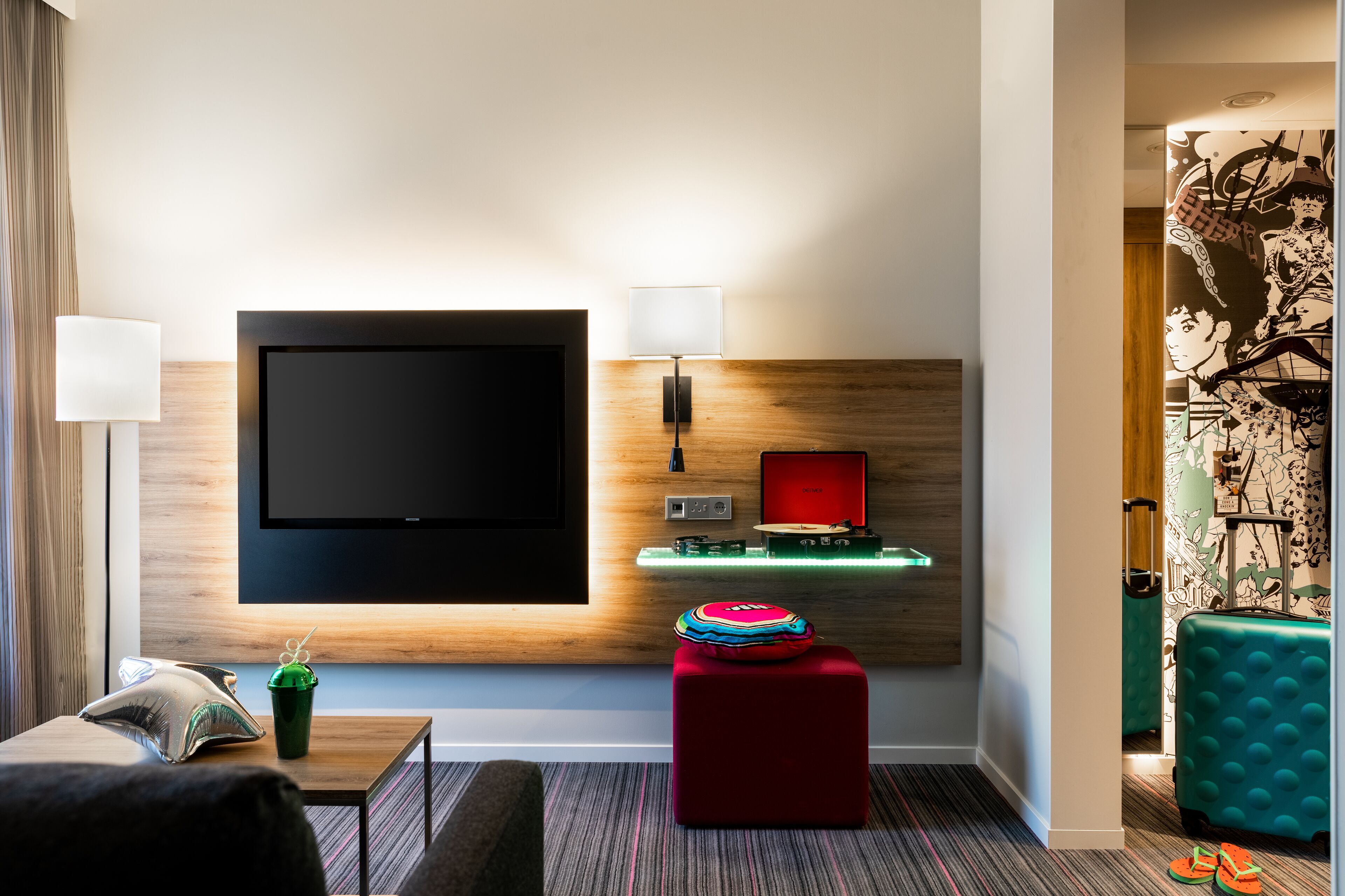 Een moderne hotelkamer met een grote flatscreen televisie aan de muur, een klein tafeltje met een lamp, een rode poef, een groene koffer en een blauwgroene koffer.