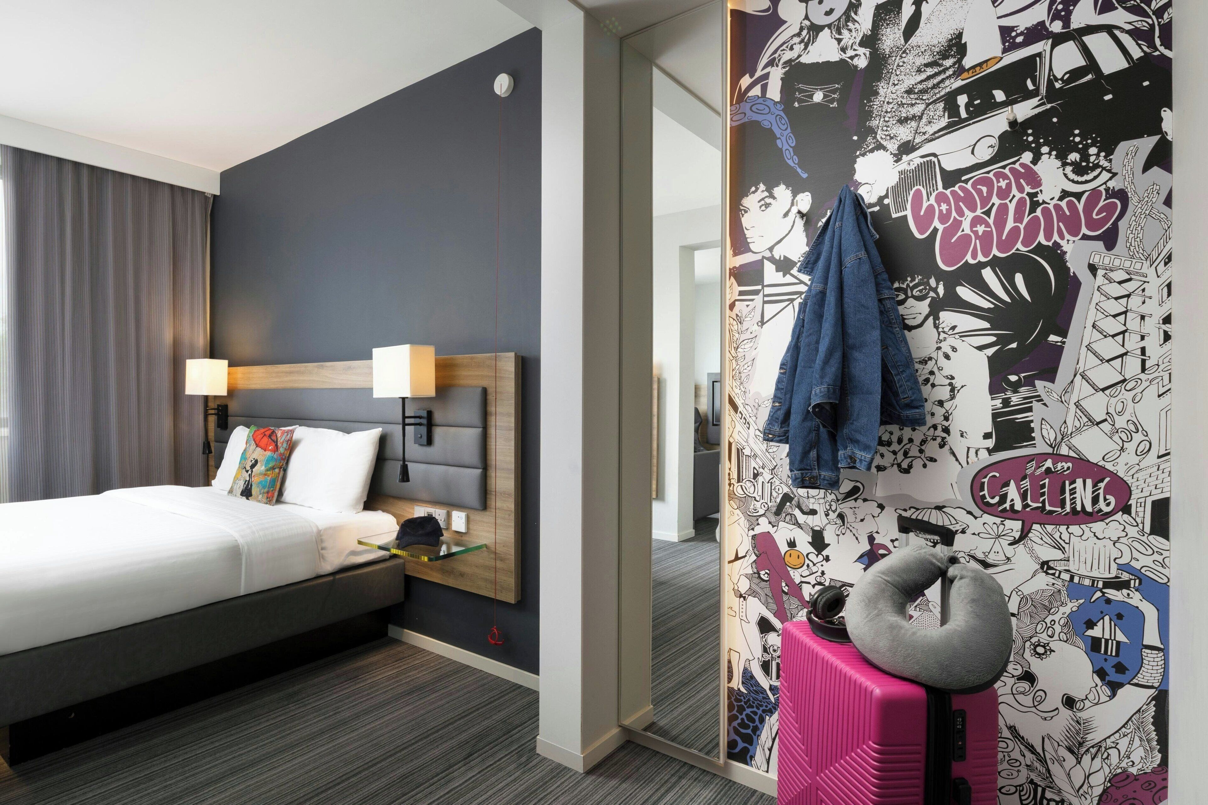 A hotel room with a bed, a desk, a lamp, a suitcase, and a wall with a London-themed mural.