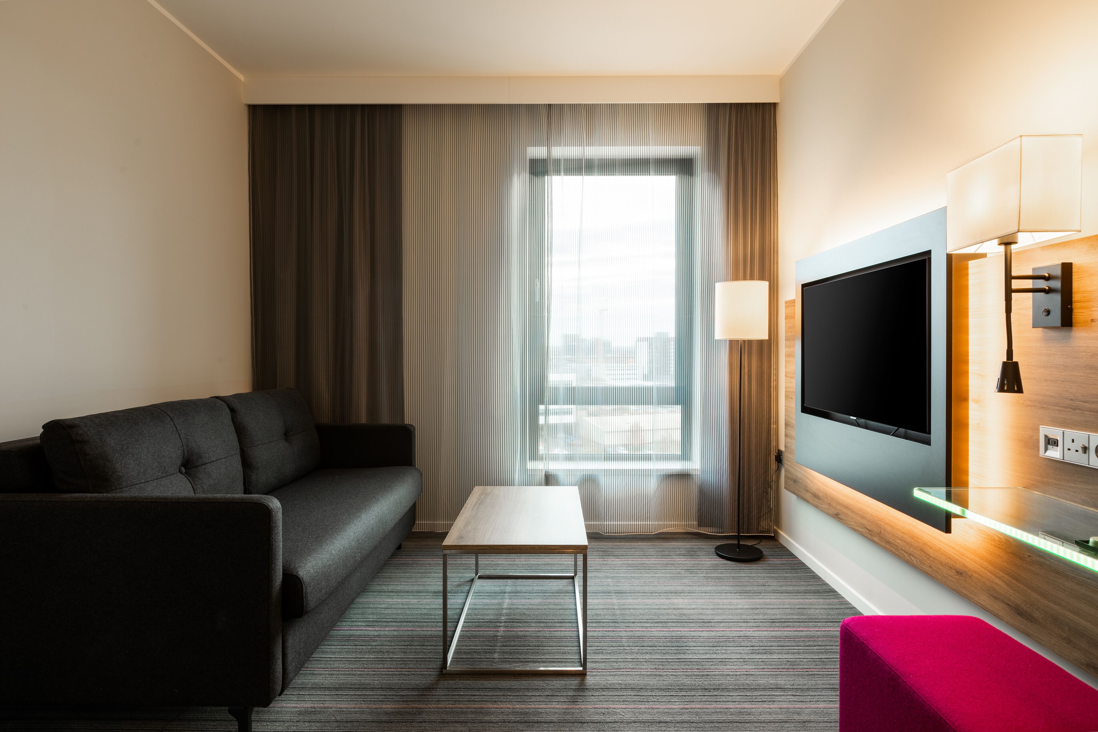 Une chambre d’hôtel avec un canapé, une petite table, un téléviseur fixé au mur et une fenêtre avec des rideaux.