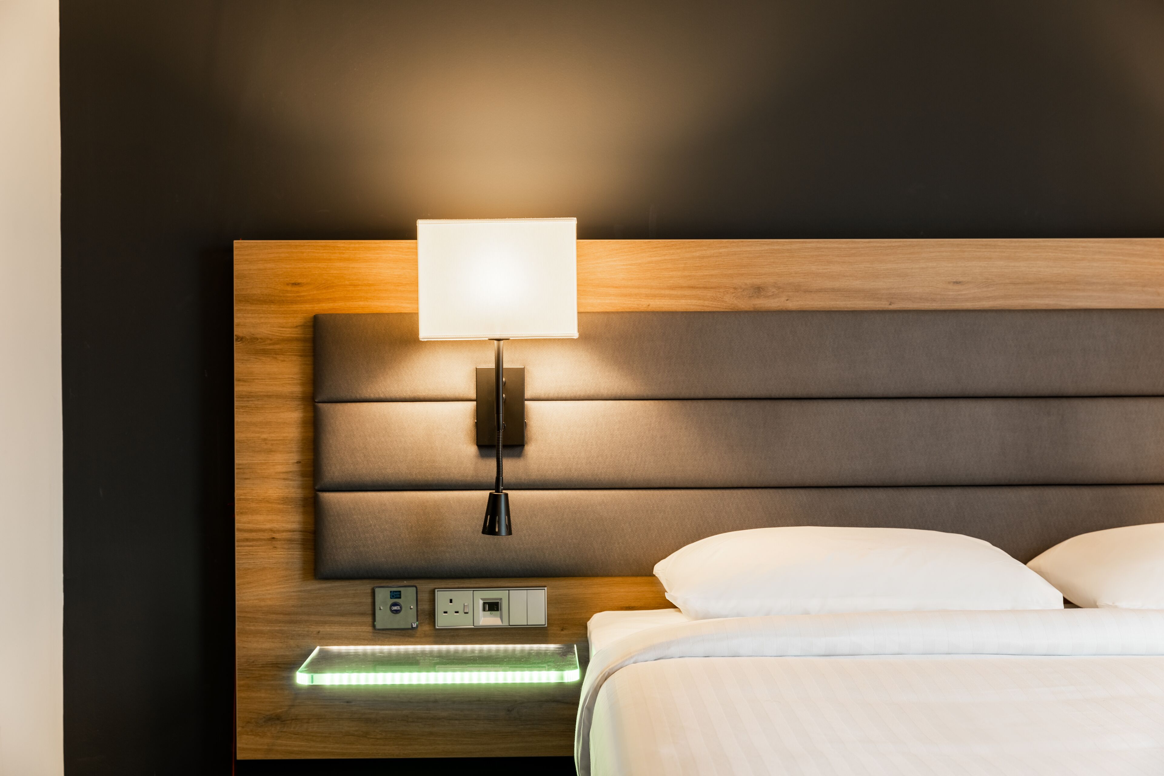 A modern hotel room with a wooden headboard, a bedside lamp, a flat-screen TV, and a power outlet.