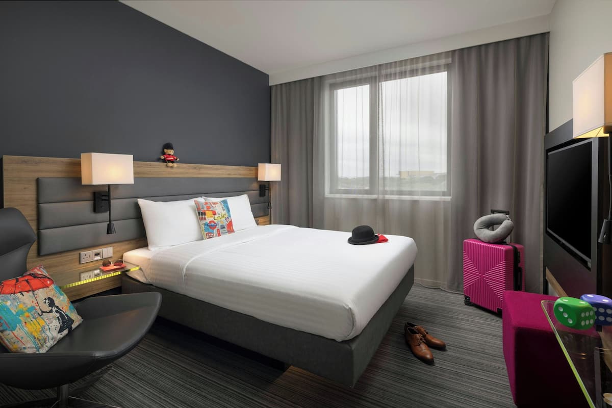 Ein Hotelzimmer mit einem großen Bett, einem Fernseher, einem Schreibtisch mit einer Lampe, einem Sessel und einem Koffer.
