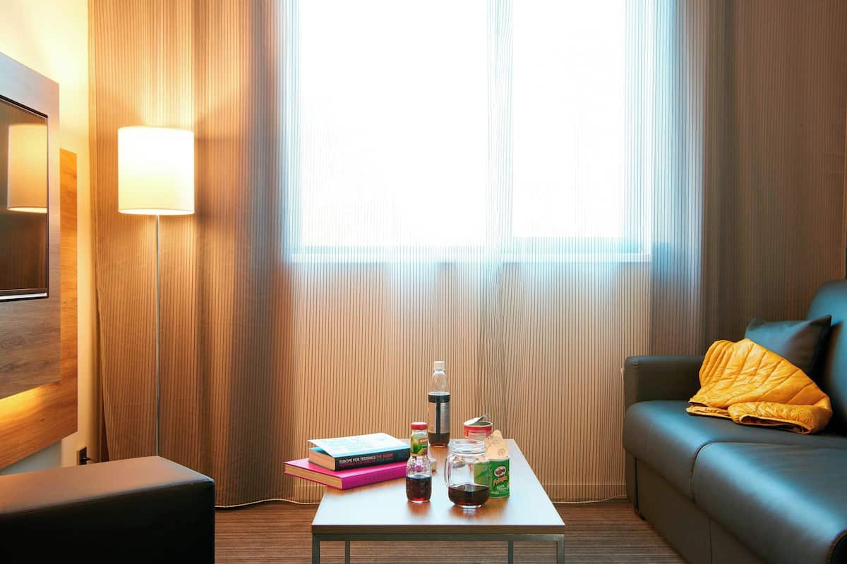 Ein modernes Hotelzimmer mit großem Fenster, einem Sofa mit gelber Decke, einem Couchtisch mit Büchern und einer Flasche sowie einer Wandlampe.