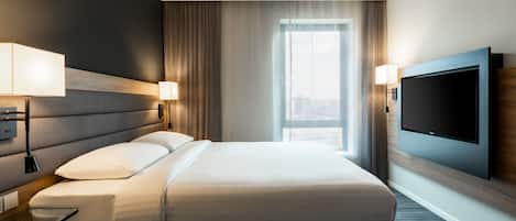 Een moderne hotelkamer met een groot bed, twee bedlampen, een flatscreen televisie aan de wand en een groot raam met gordijnen.