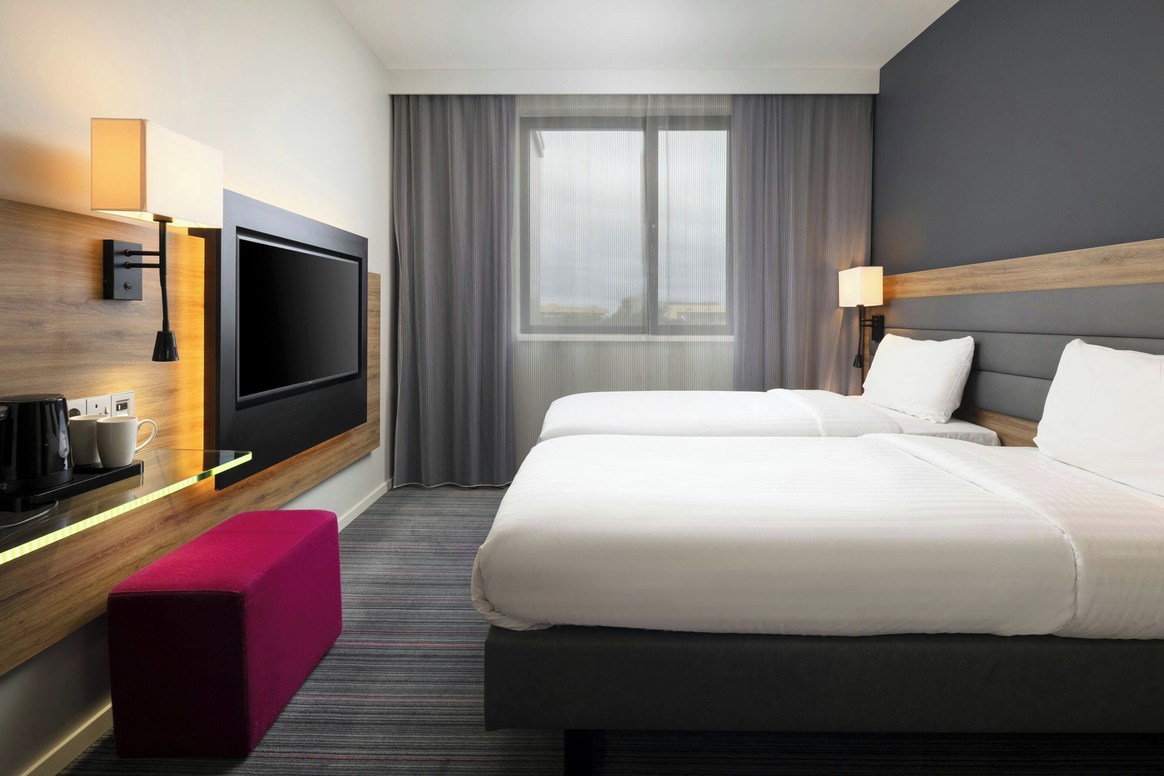 A hotel room with a large bed, a flat-screen TV, a desk with a lamp, and a window with curtains.