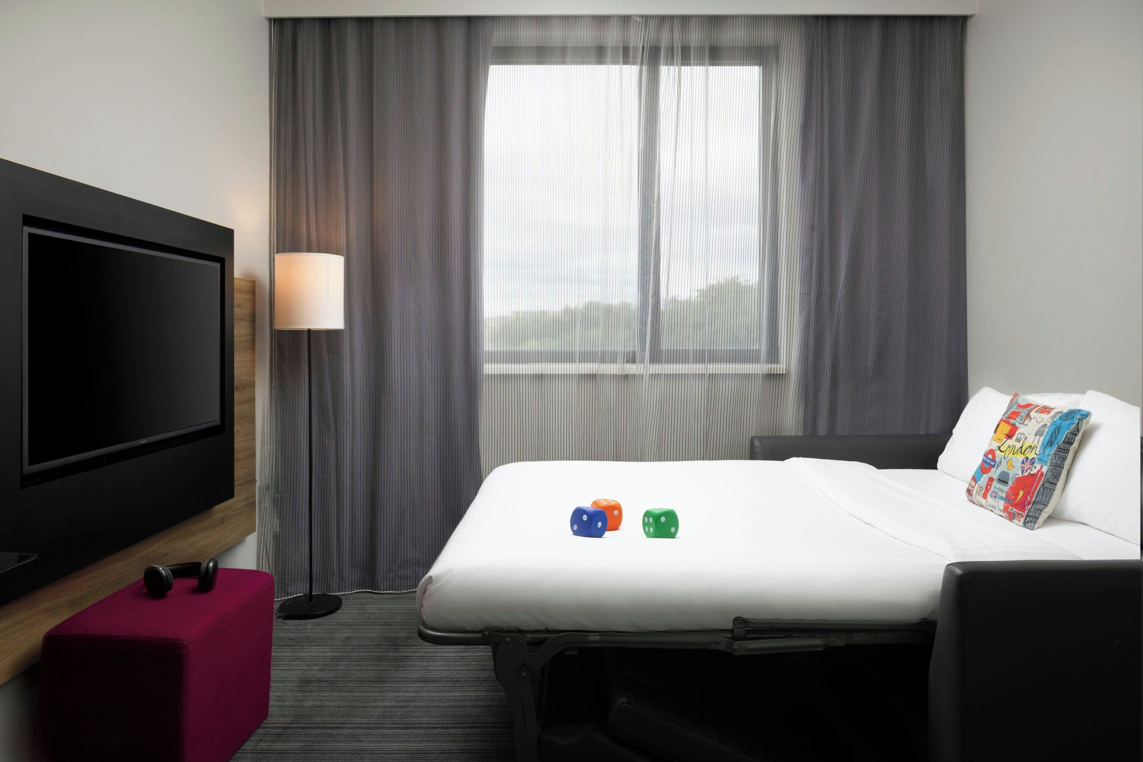Habitación de hotel con cama, televisión, lámpara y ventana con cortinas.
