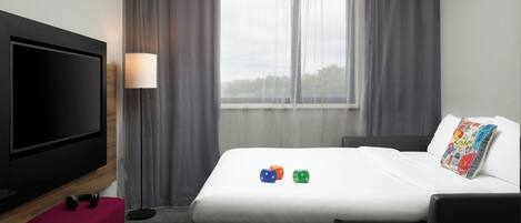 Habitación de hotel con cama, televisión, lámpara y ventana con cortinas.