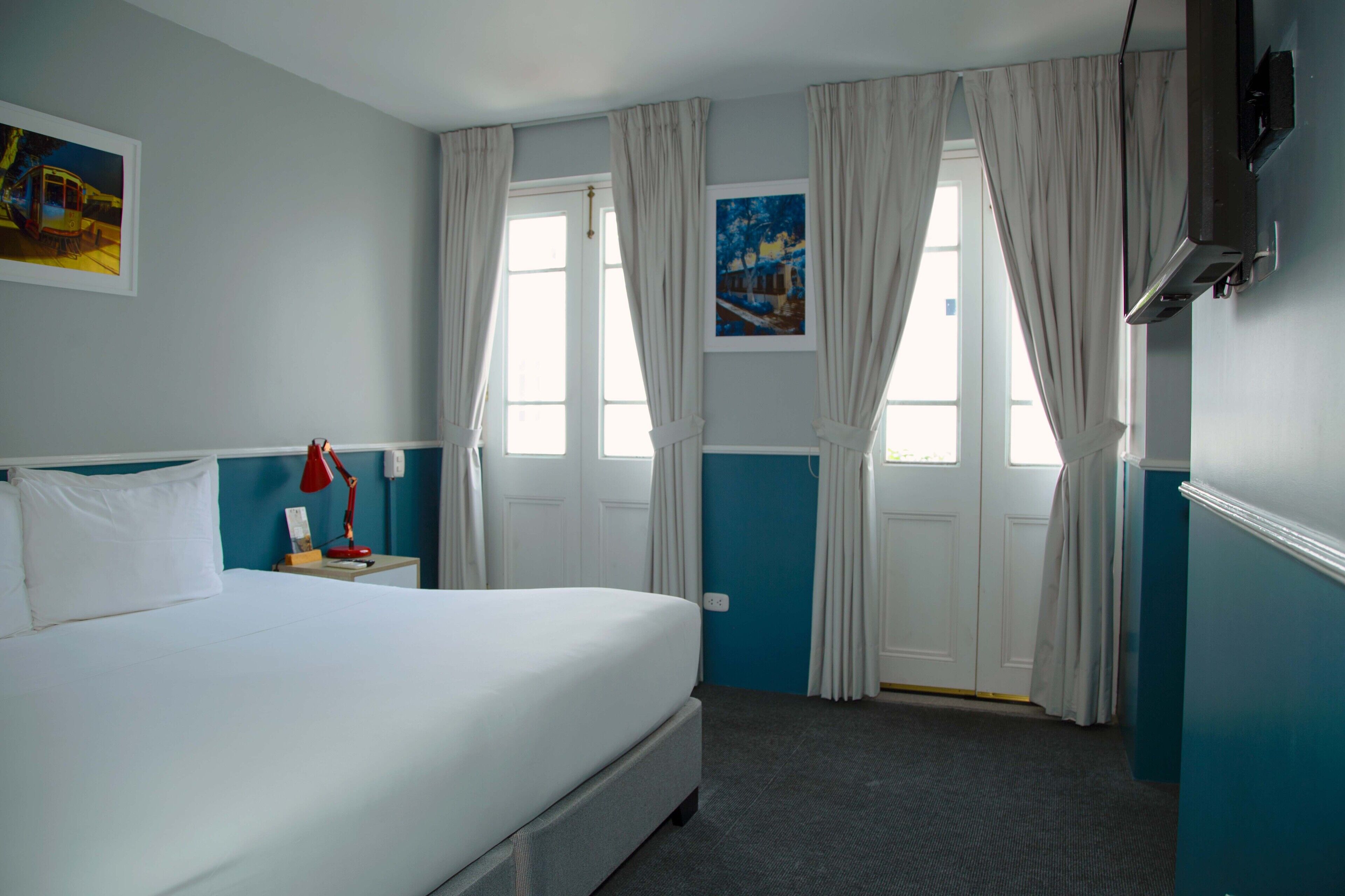 Una habitación de hotel con una cama, una mesita de noche con lámpara y dos ventanas con cortinas.