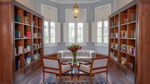 Uma biblioteca com estantes de madeira, uma área de estar central com um sofá e cadeiras, um vaso com flores e um candelabro.