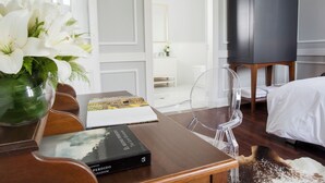 Quarto de hotel com escrivaninha de madeira, um vaso de flores brancas, um livro, uma cadeira transparente, uma cama e um armário escuro.
