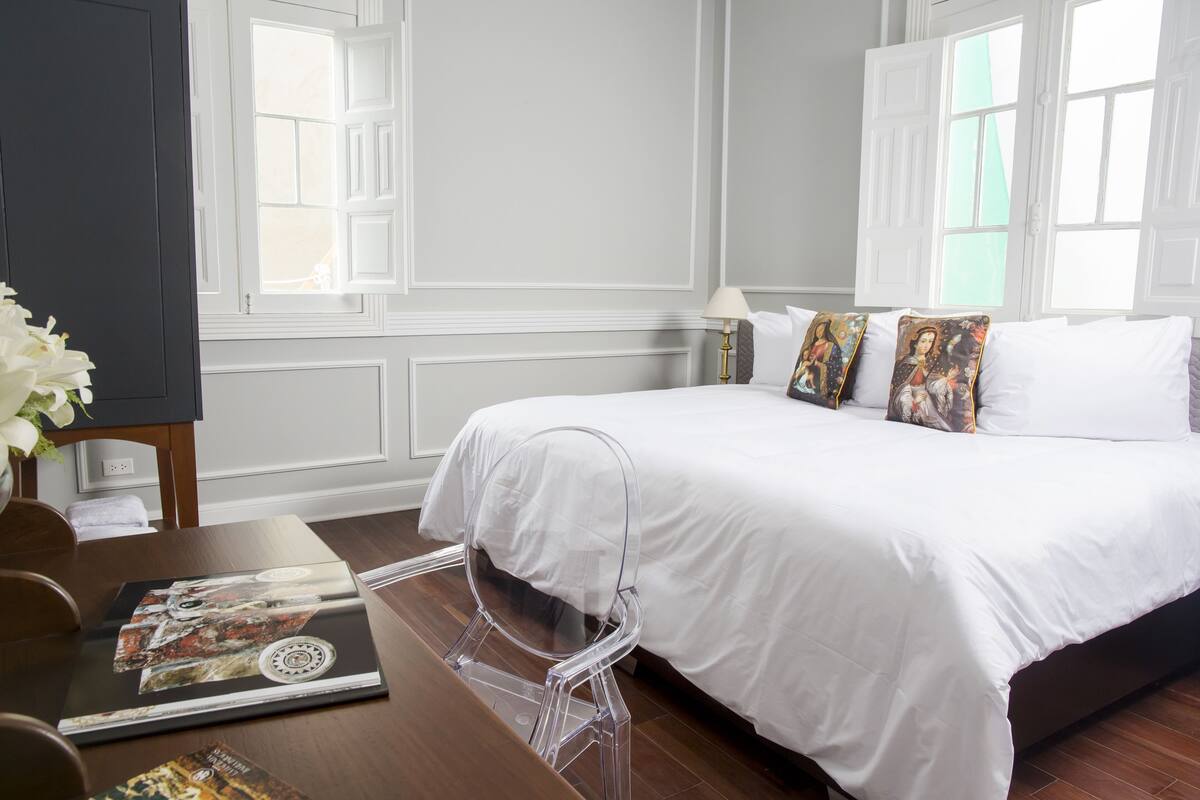 A hotel room with a large bed, white bedding, and decorative pillows. There is a clear plastic chair and a table with magazines.