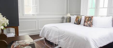 A hotel room with a large bed, white bedding, and decorative pillows. There is a clear plastic chair and a table with magazines.