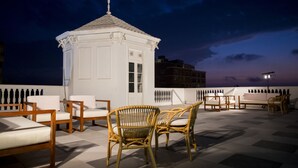 Terraço no telhado com mobiliário de vime, um pequeno gazebo branco e vista para a cidade ao entardecer.