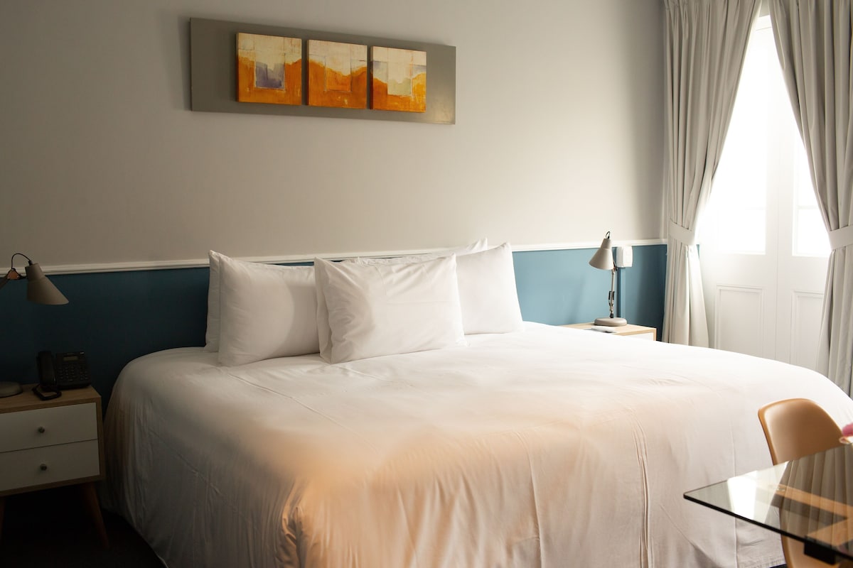 A hotel room with a bed, bedside table, lamp, and a painting on the wall.