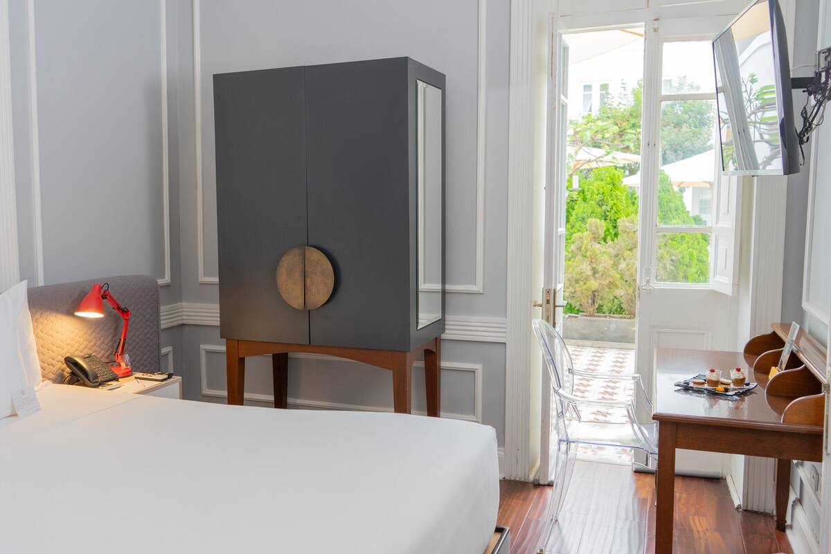 A hotel room with a bed, a desk, a wardrobe, a desk lamp, and a window with a view of greenery.