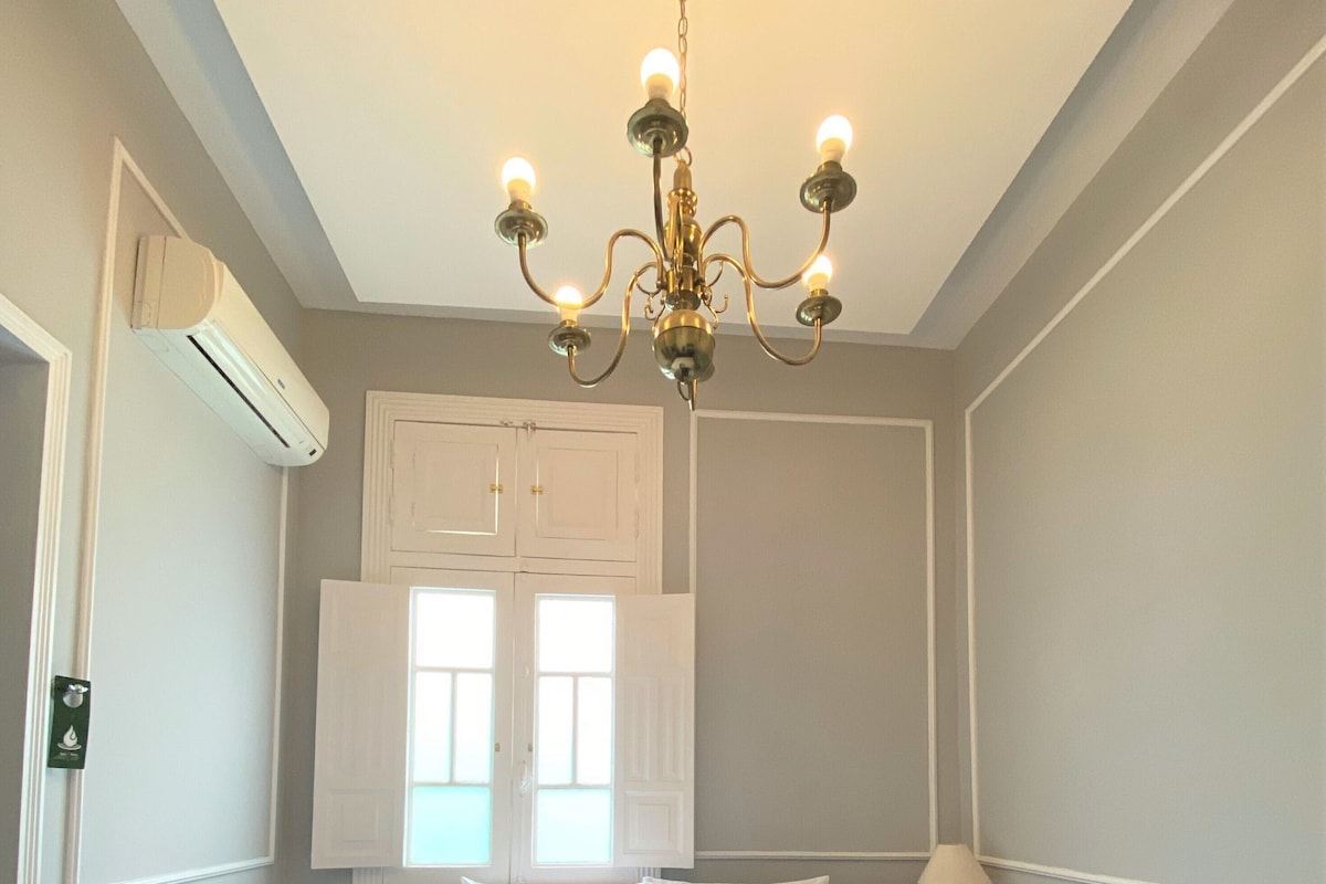 A bedroom with a large bed, a chandelier, a window with shutters, and an air conditioning unit.