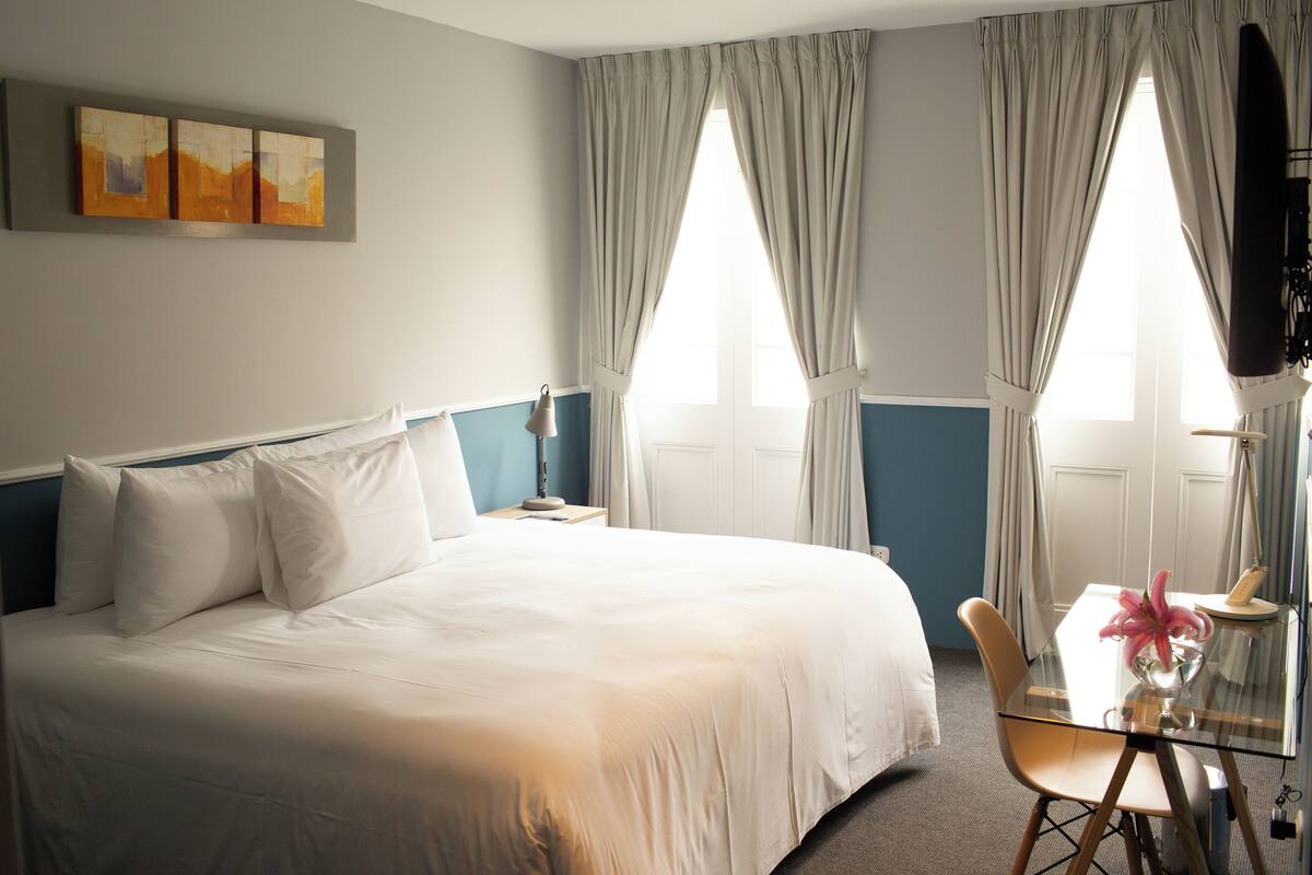 A hotel room with a large bed, a desk with a chair, and two windows with curtains.