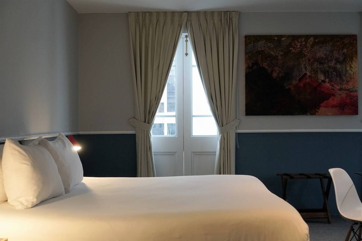 A hotel room with a bed, a chair, a table, a window with curtains, and a painting on the wall.