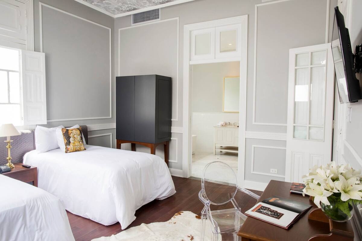 A hotel room with a bed, a TV, a desk with a vase, and a bathroom visible through an open door.