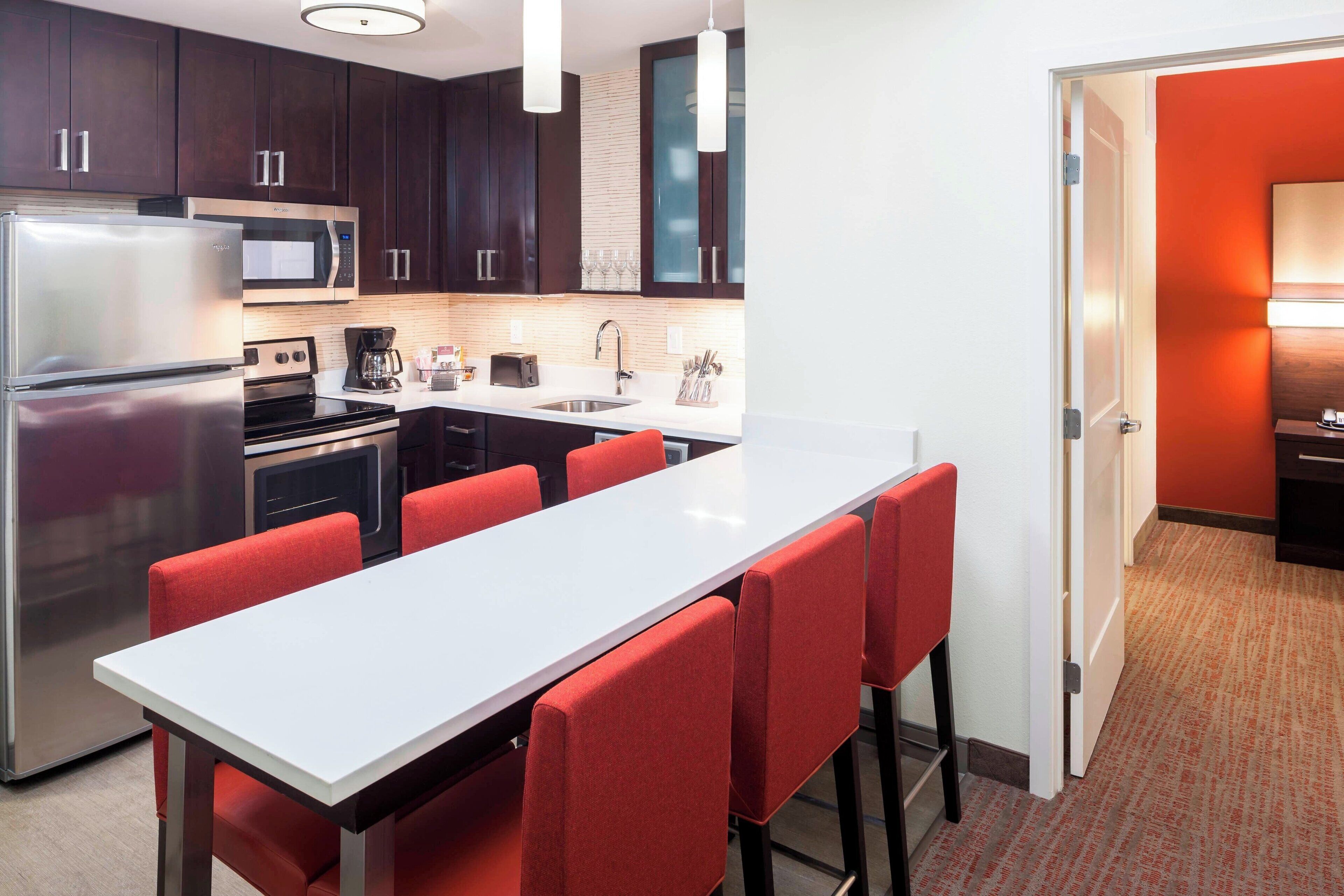 A modern kitchen with dark wood cabinets, stainless steel appliances, and a white countertop. There are red chairs around a central island.