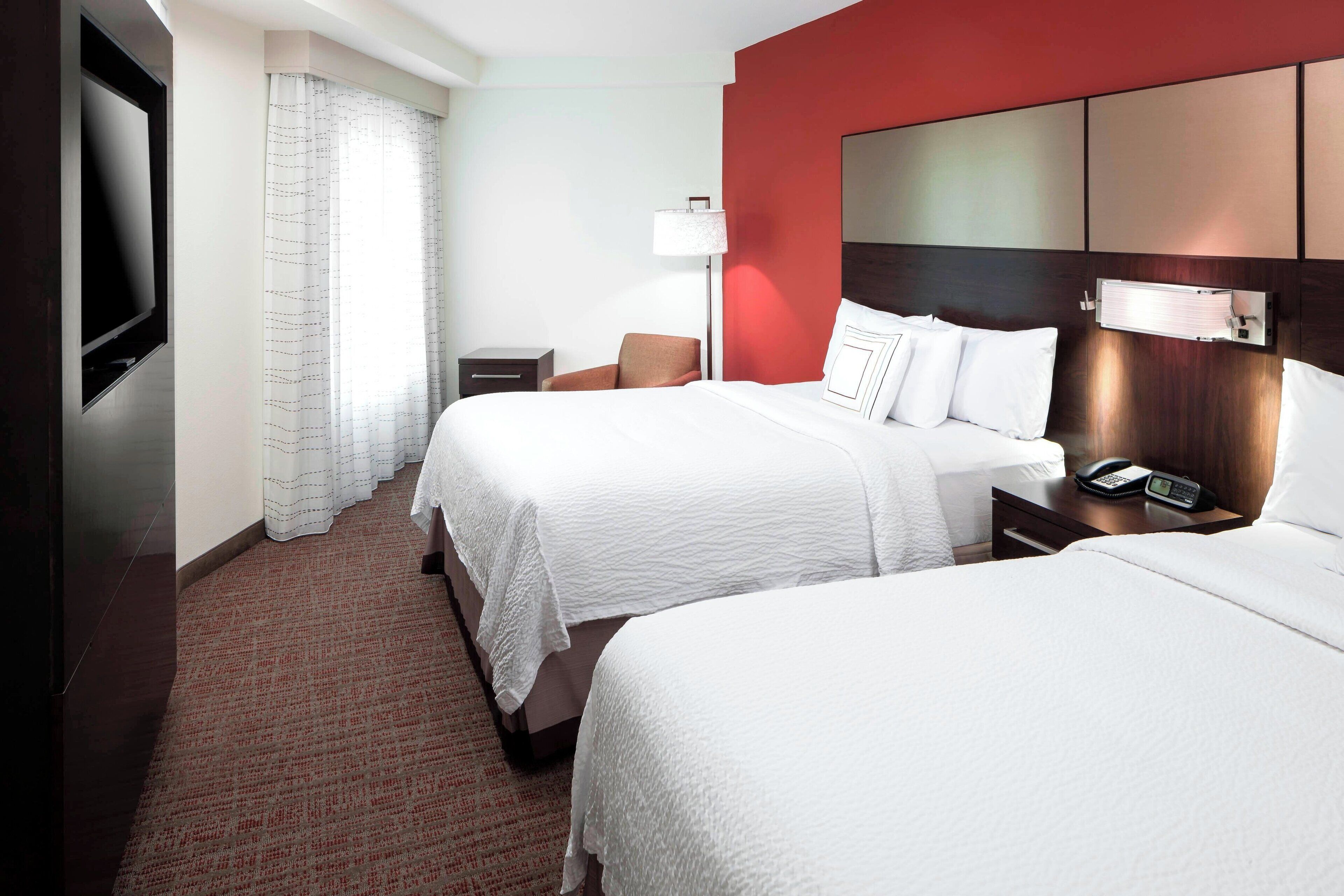 A hotel room with two beds, a TV, a bedside table with a lamp, and a red accent wall.