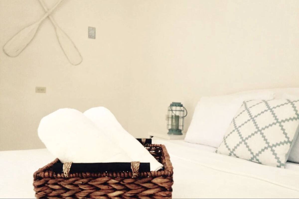 A neatly made bed with a white pillow and a patterned pillow, a wicker basket with a white towel, and a wall-mounted light fixture.