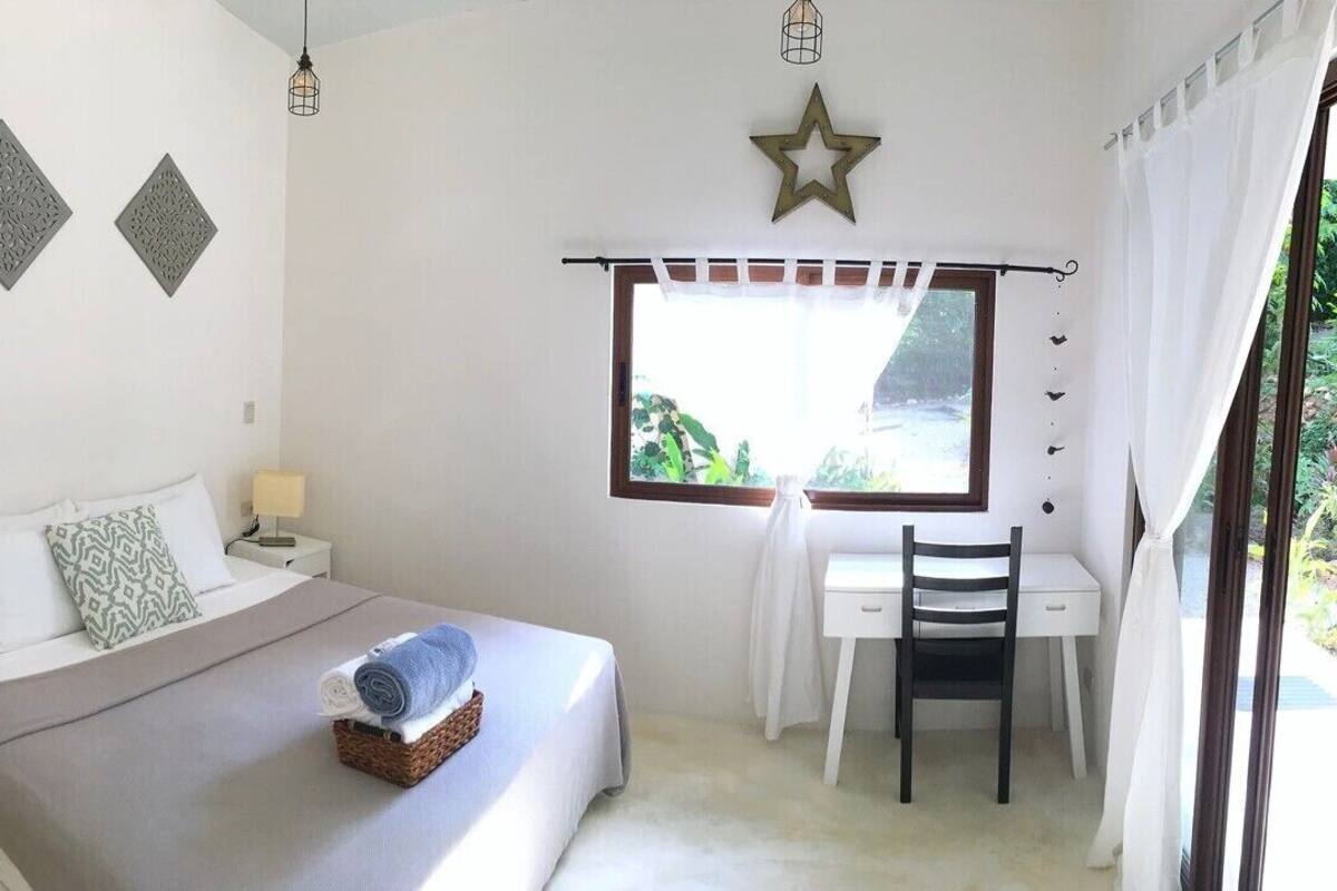 A bedroom with a bed, bedside table, and a desk with a chair. There is a window with curtains and a star decoration on the wall.