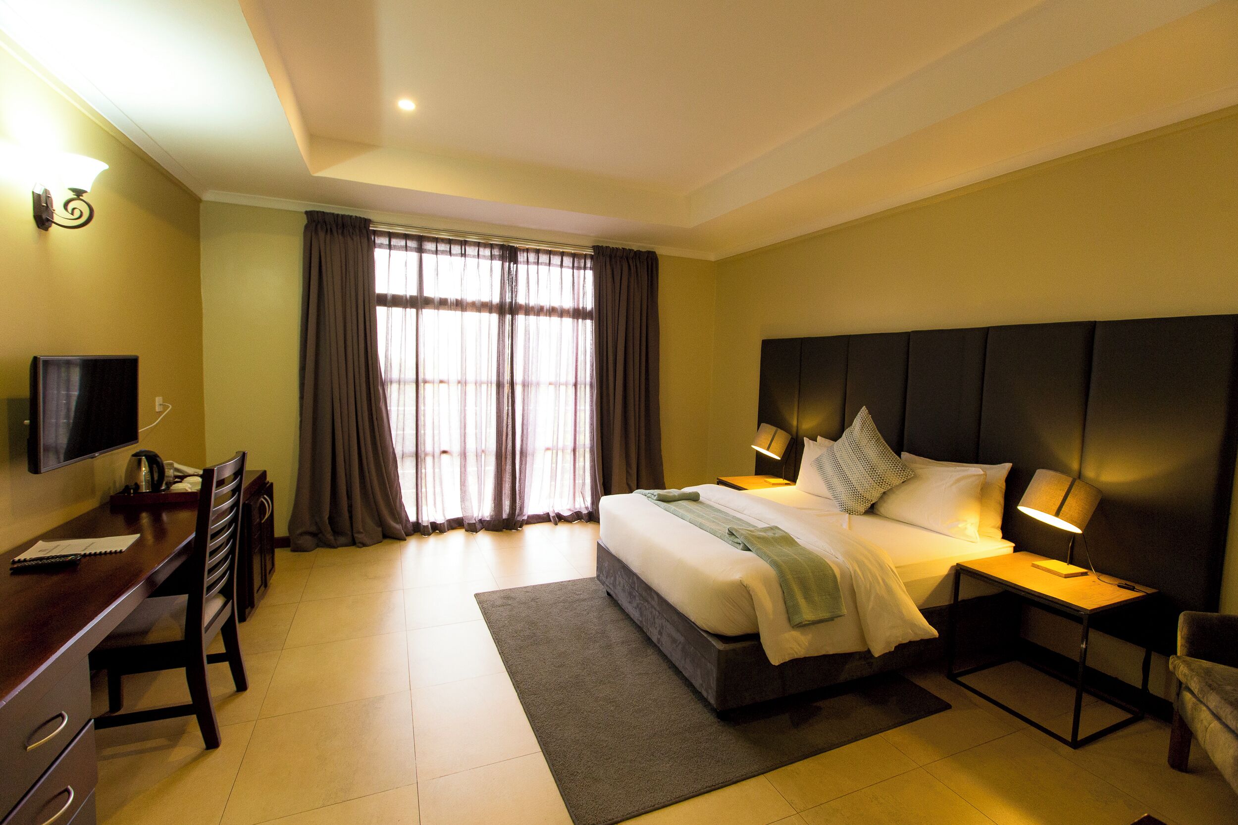 Une chambre d’hôtel comprenant un lit, une table de chevet, une lampe, un bureau avec une chaise, une télévision et des rideaux.