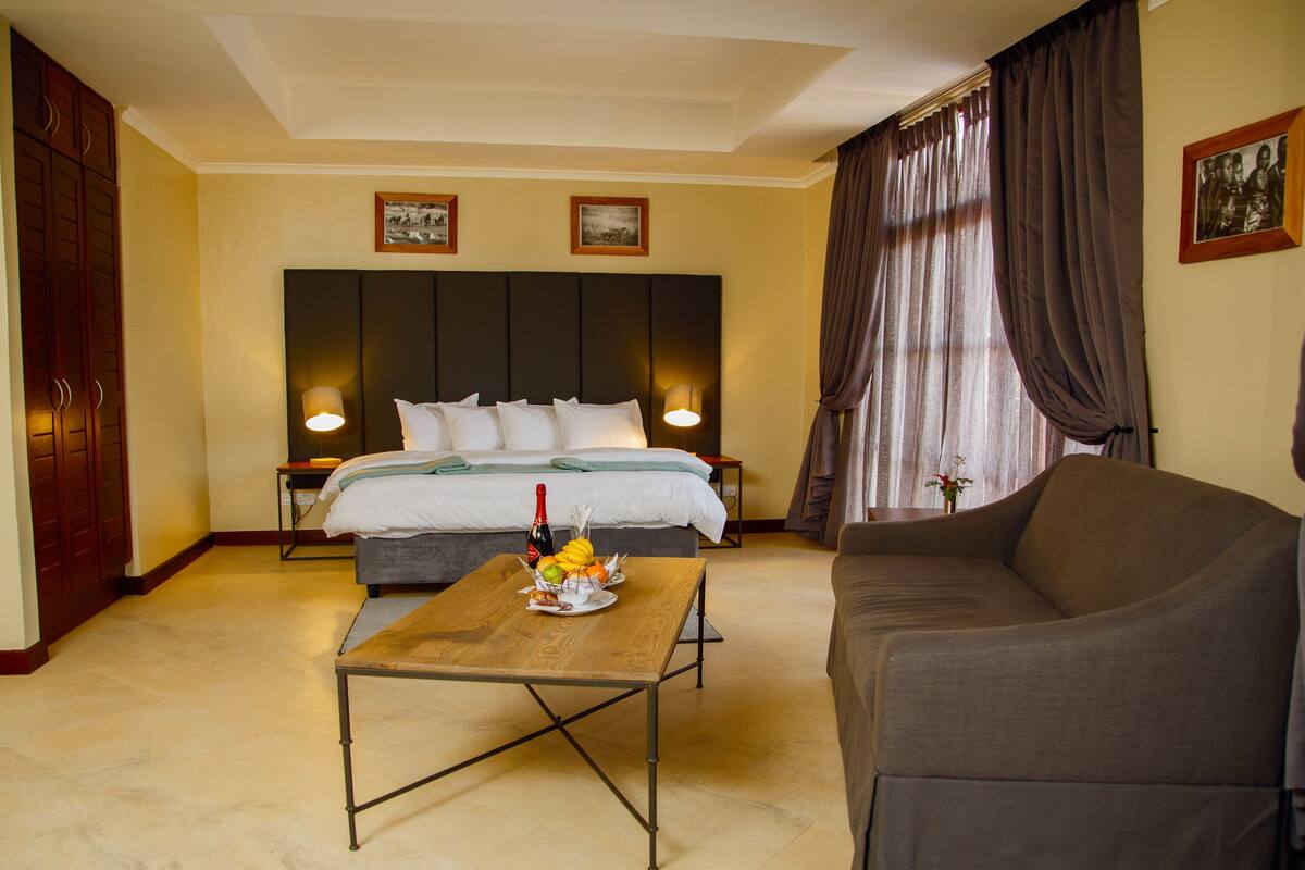 Een hotelkamer met een bed, een bank, een tafel met fruit en een fles, en gordijnen.