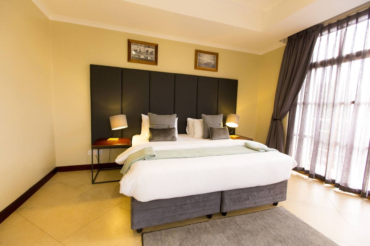 Een hotelkamer met een groot bed, twee nachtkastjes, een hoofdeinde en uitzicht door dunne gordijnen.
