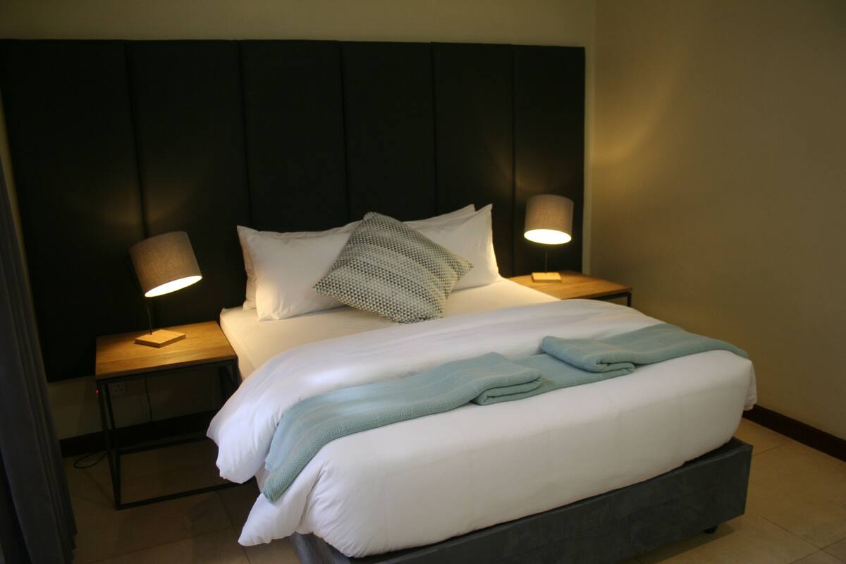 Een netjes opgemaakt bed met witte lakens en een lichtblauwe plaid, met aan beide zijden nachtlampjes op houten nachtkastjes.