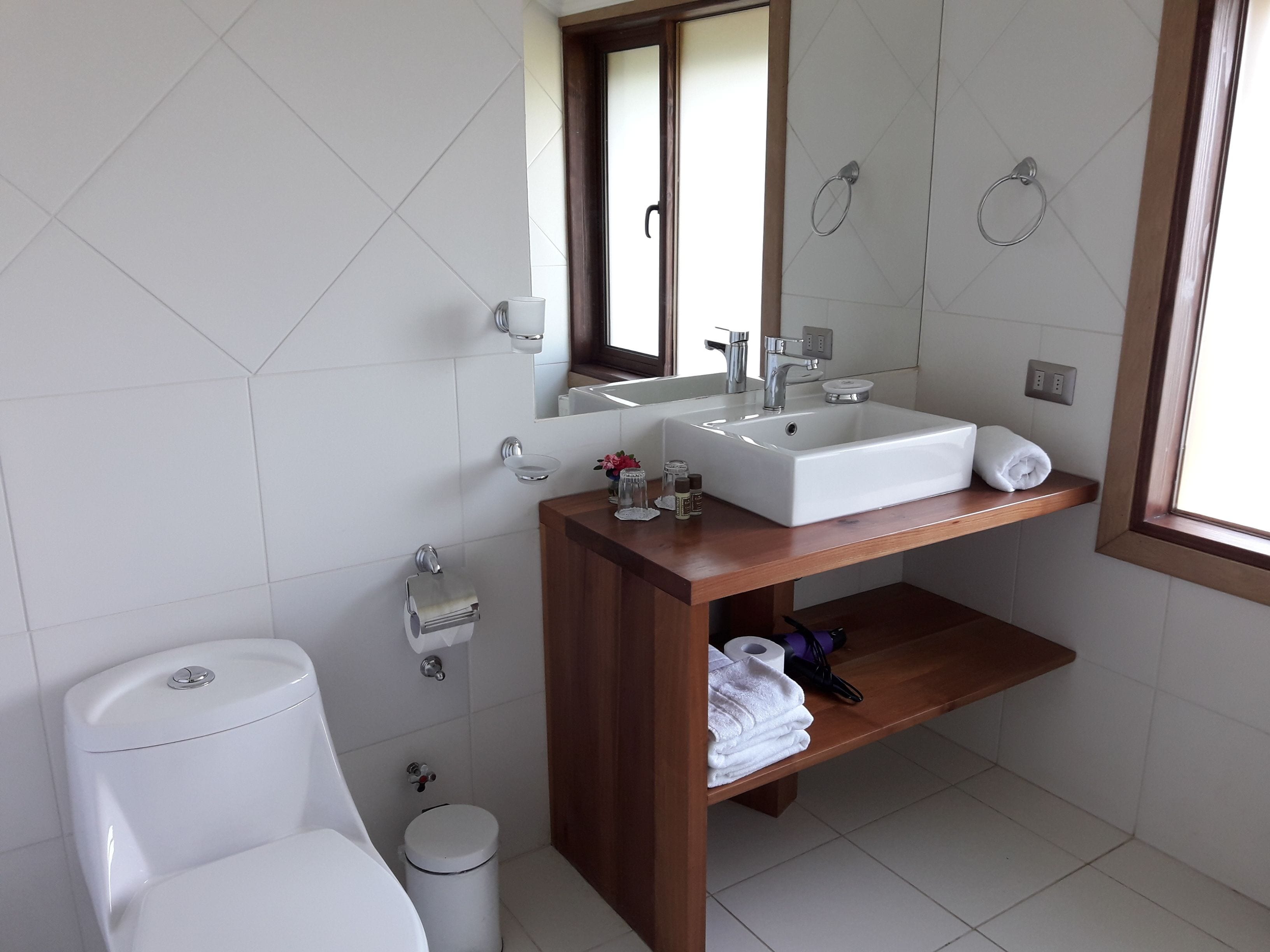Un baño moderno con inodoro blanco, lavamanos con espejo encima y repisa de madera debajo.