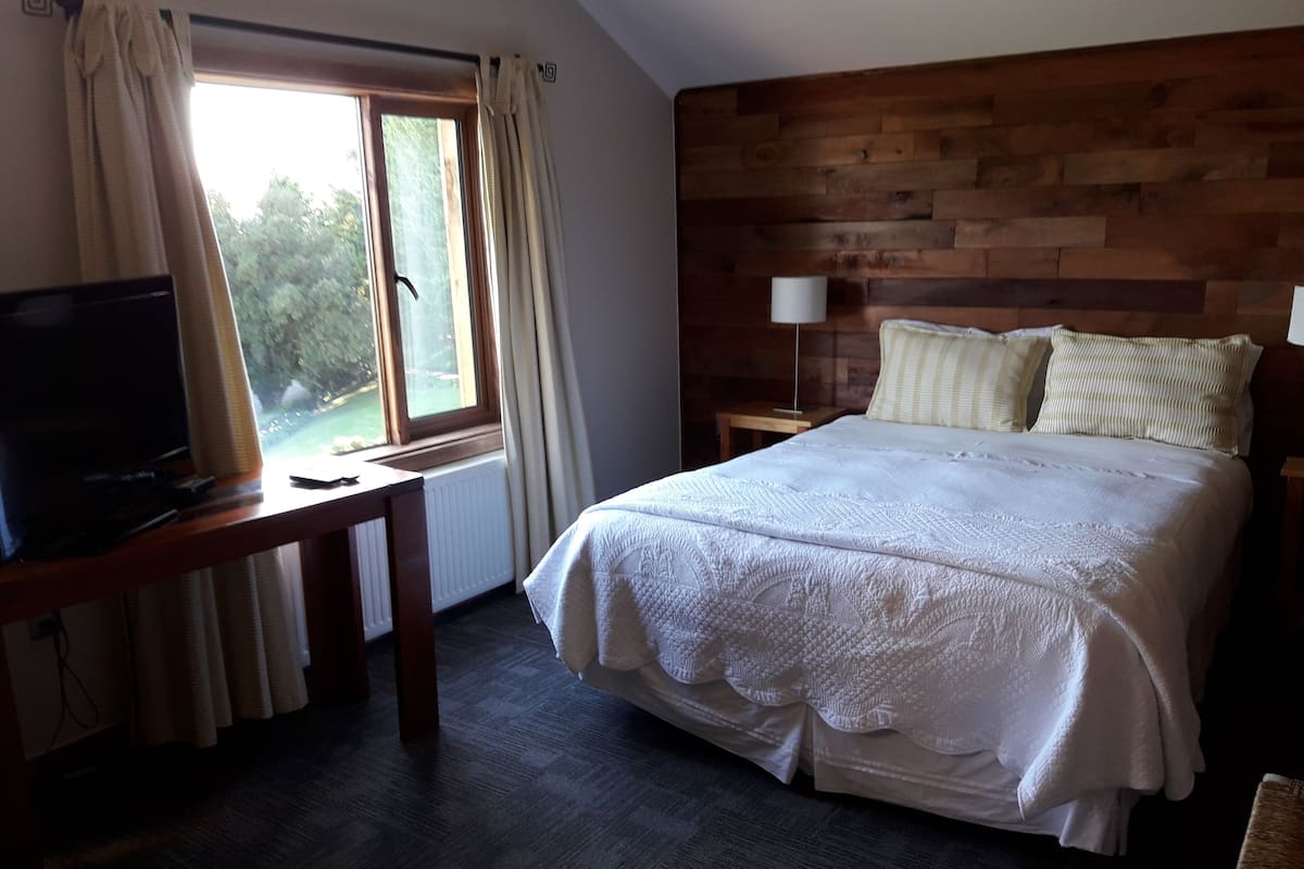 Un dormitorio con cabecera de madera, cama con ropa de cama blanca, mesita de noche con lámpara y televisor sobre un mueble de madera.