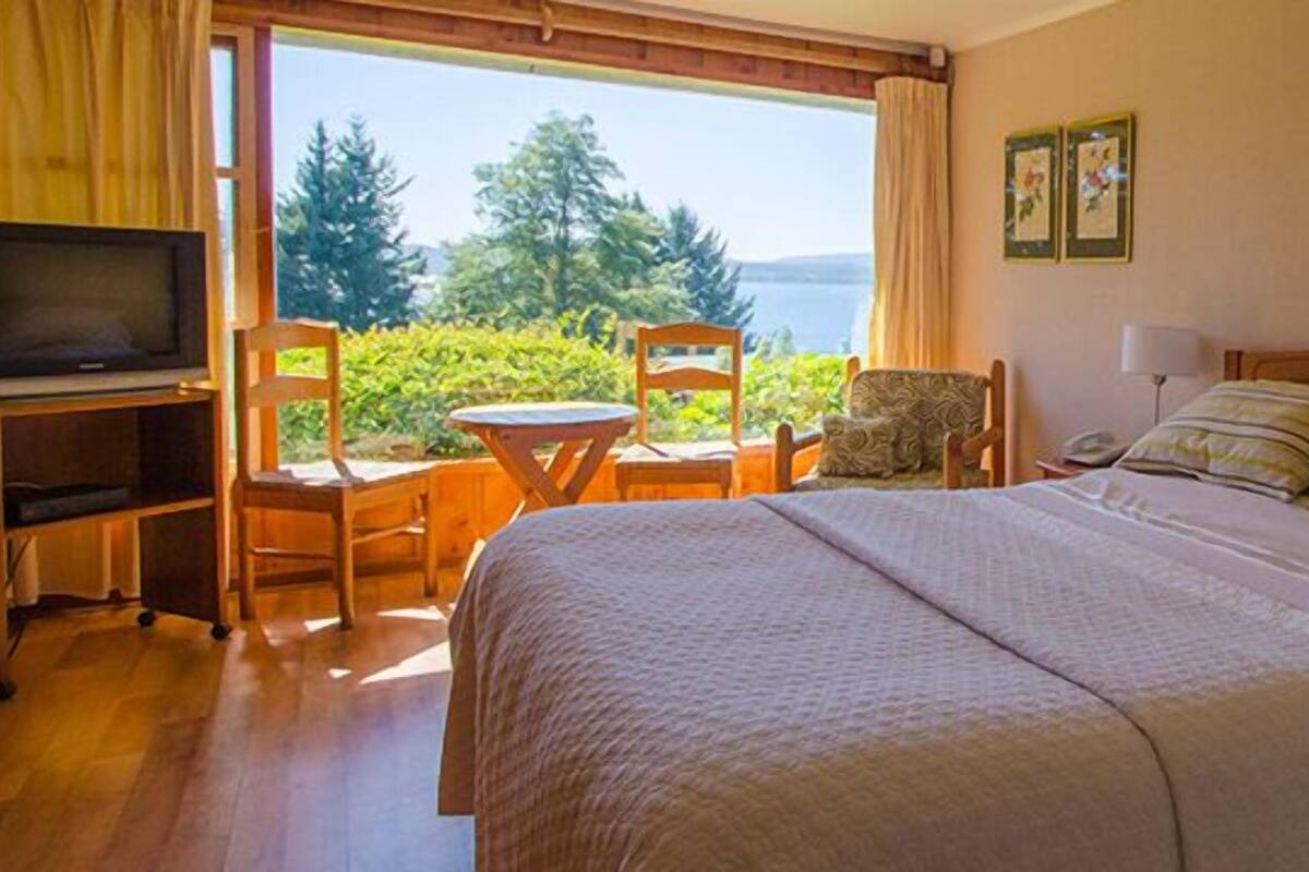 Habitación de hotel con cama, televisor, una mesita y una silla. Se ve una ventana con vistas a árboles y un cuerpo de agua.