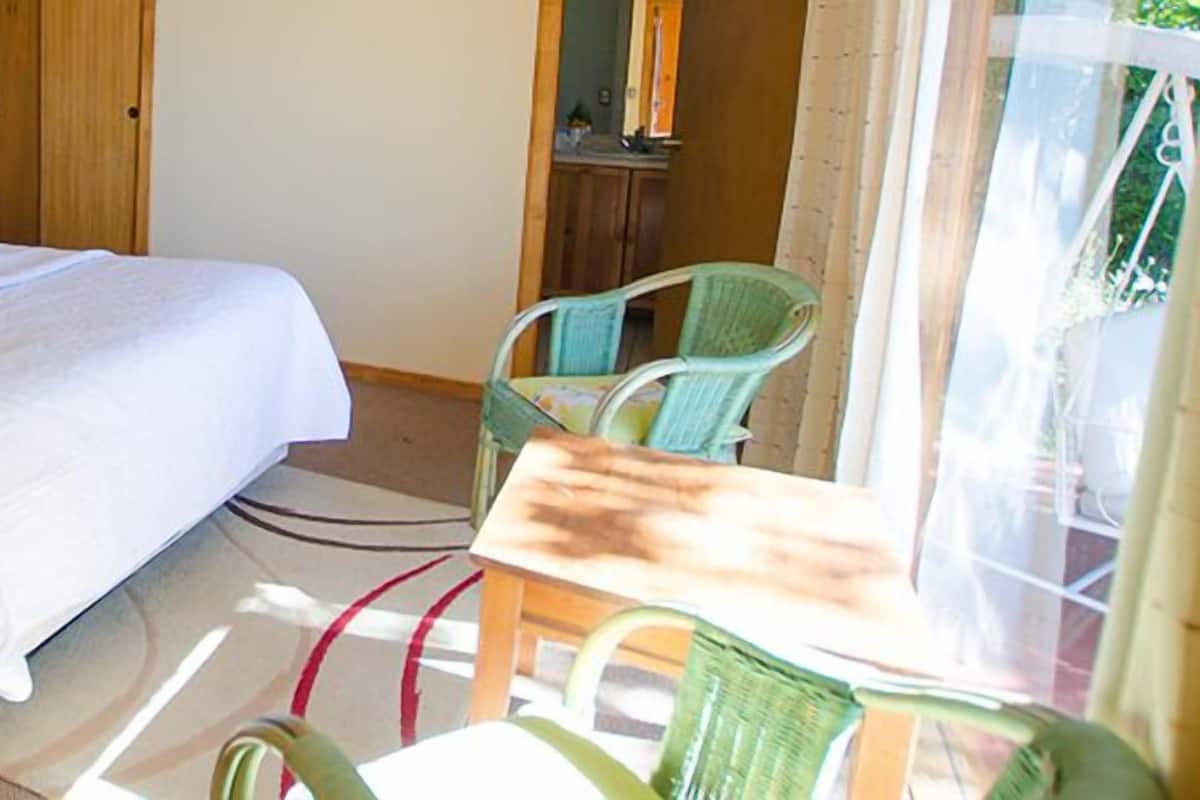 Un dormitorio con cabecera de madera, cama con ropa de cama blanca, dos lámparas de noche, una silla verde y vistas al exterior.