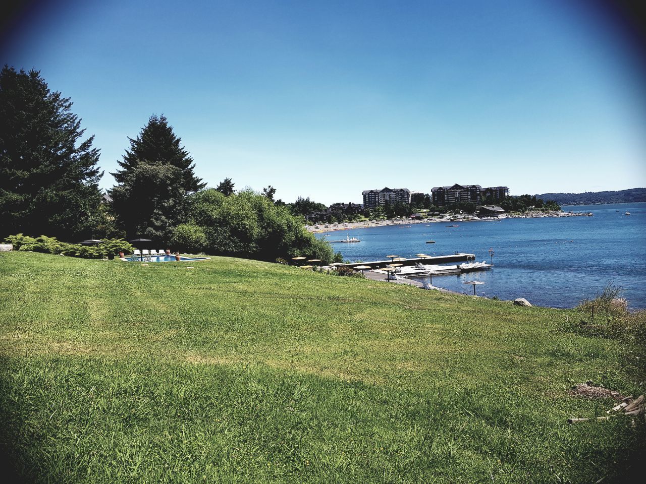 A grassy area with a dock and boats, a clear blue sky, and buildings in the background.