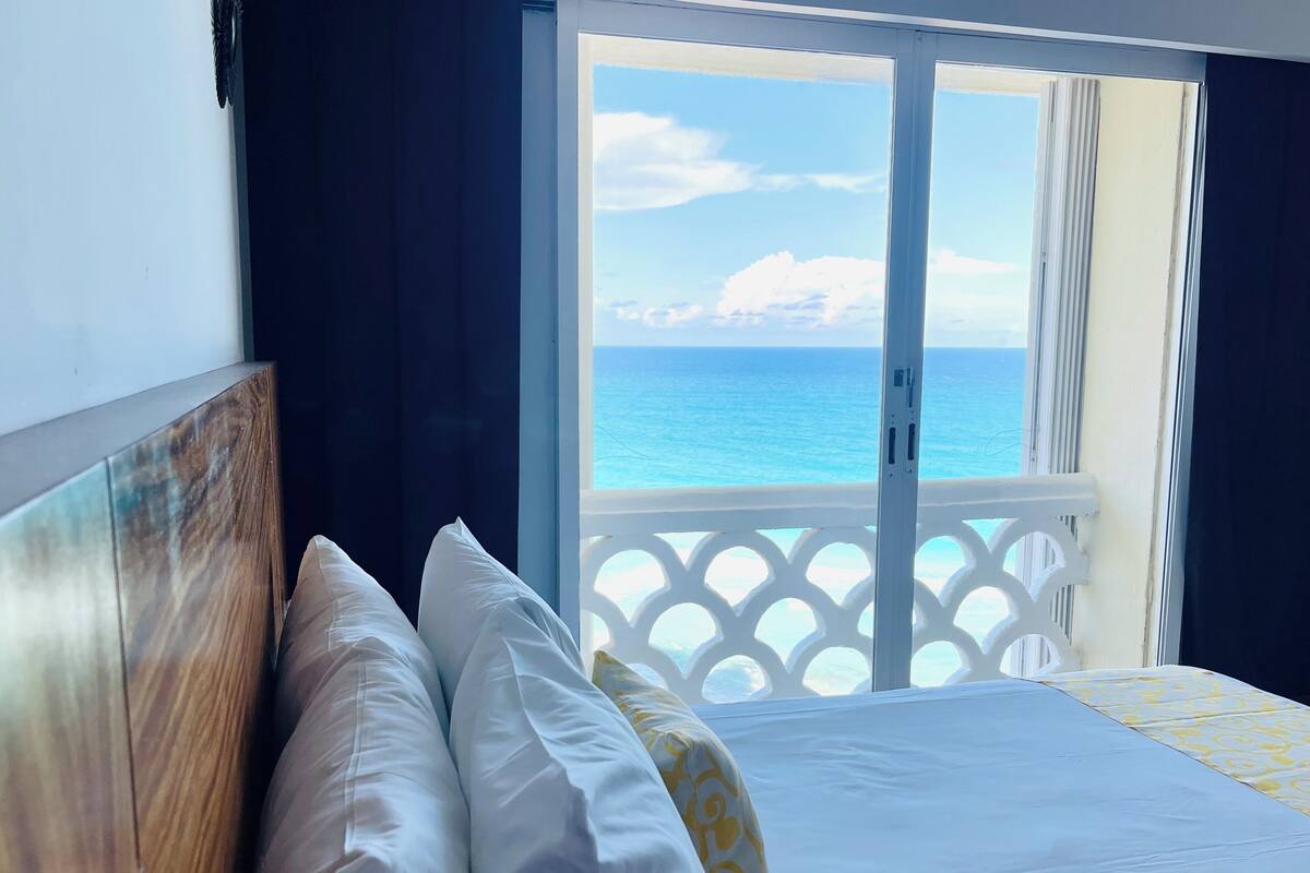 Una habitación con una cama, vista al océano desde un balcón y barandilla blanca.