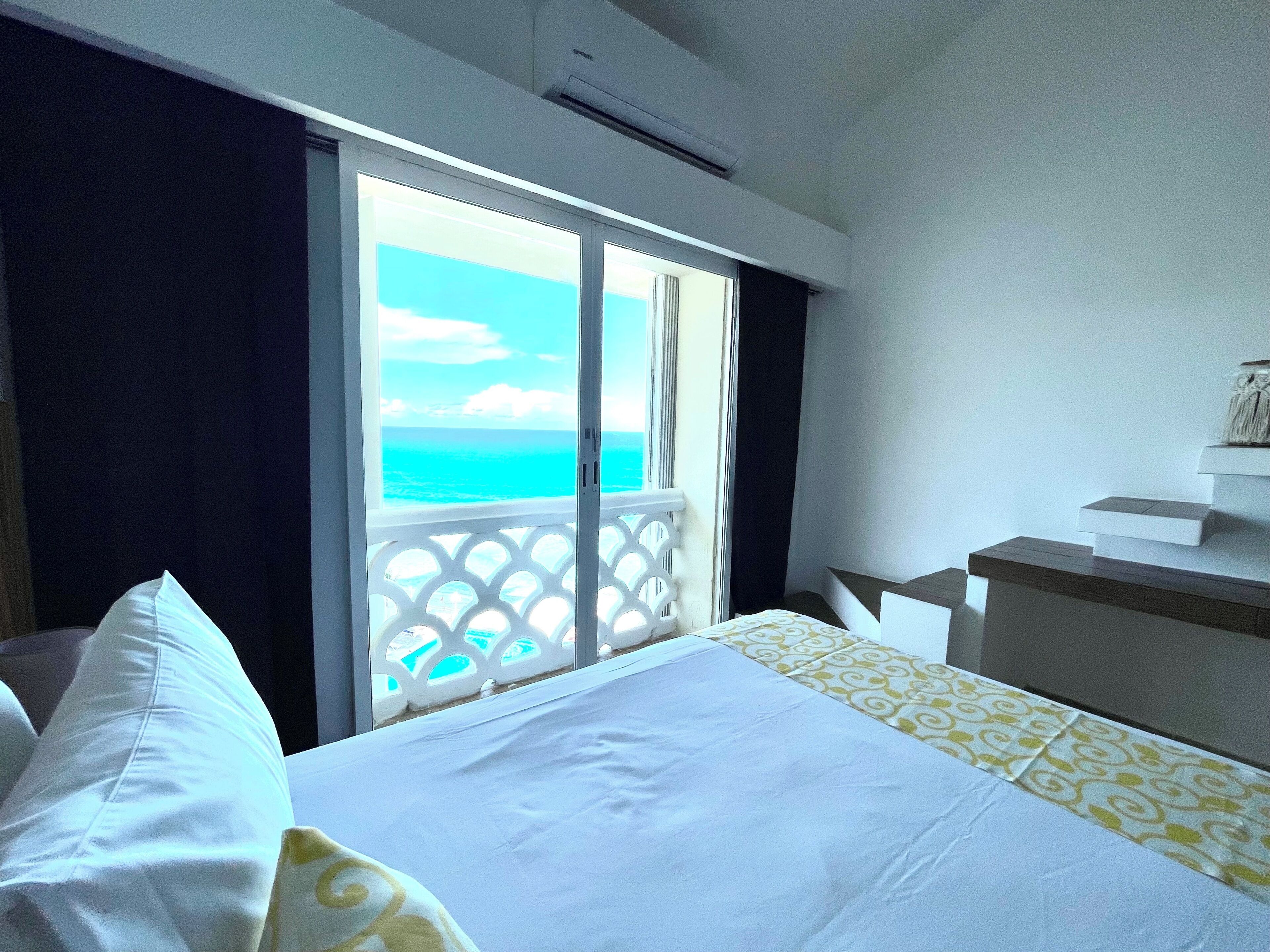 A hotel room with a bed, a small desk, and a balcony with a view of the ocean.