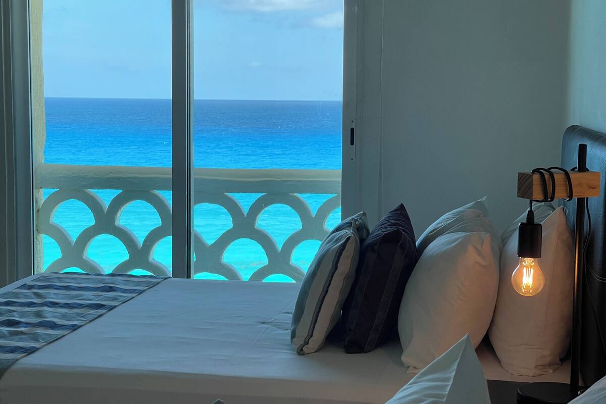 Una cama con almohadas y una mesita de noche con lámpara, con vista a un mar turquesa a través de un balcón con barandilla blanca.