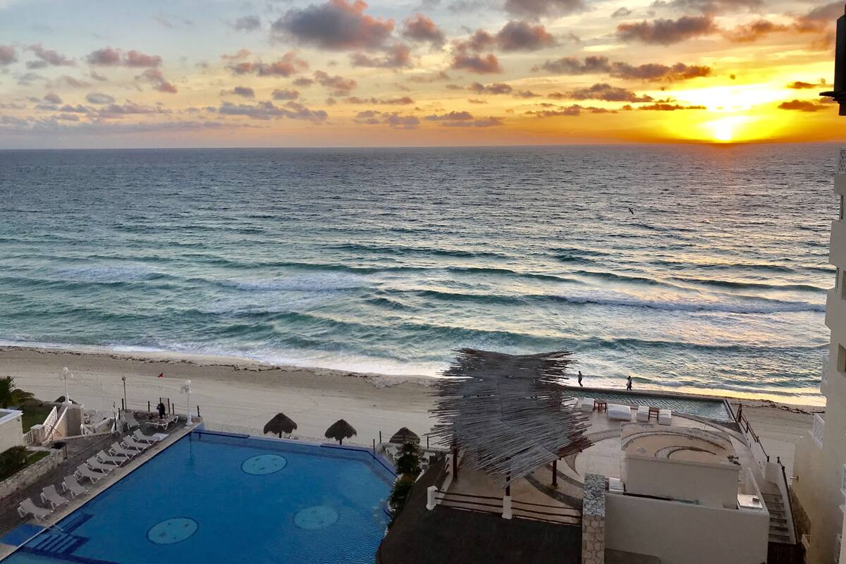 Vista a la piscina con un atardecer sobre el océano, una playa y un edificio con piscina y sombrillas.