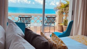A hotel room with a large window overlooking a beach, a bed with white and patterned pillows, and a table with chairs outside. - Ocean View Apartments (Cancun)