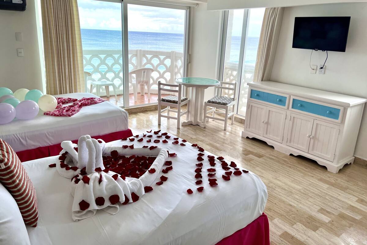 Habitación de hotel con cama, televisión, balcón con vista al mar y zona de comedor con sillas y mesa.