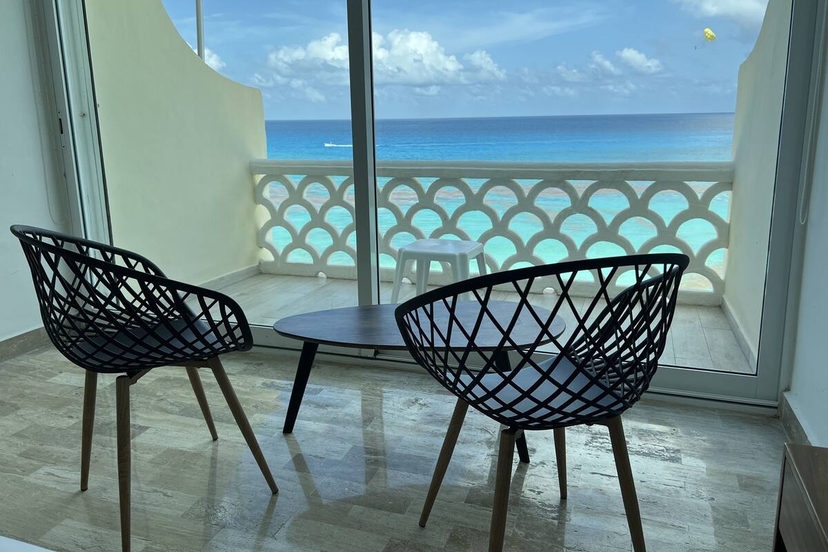 Un balcón con dos sillas de estructura metálica negra y una mesita, con vistas al mar turquesa y un cielo despejado.