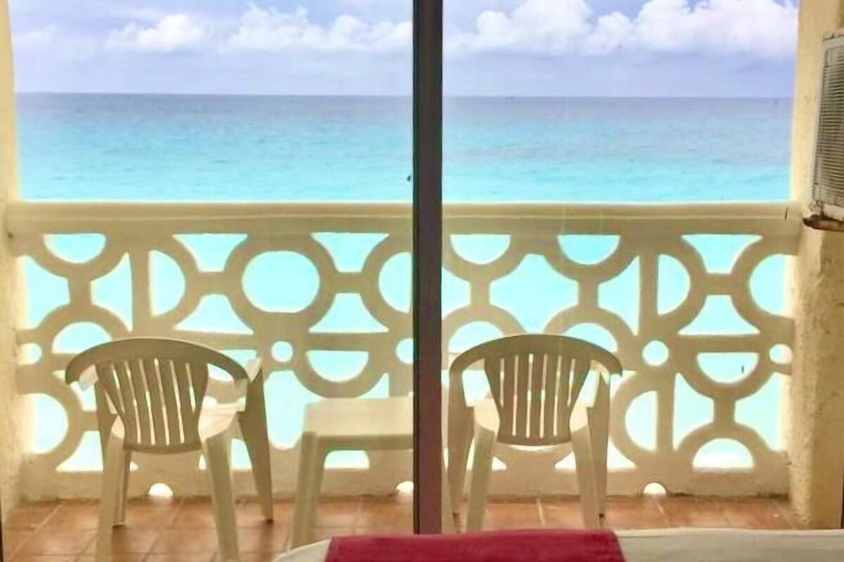 Un balcón con dos sillas y una cama con una colcha roja, con vista al mar turquesa.