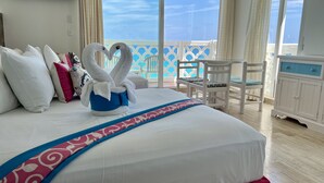 A hotel room with a large bed, a blue and white patterned bed runner, white pillows, a white swan-shaped towel arrangement, a white chair, and a view of the ocean through large windows. - Ocean View Apartments – Increíble (Cancun)