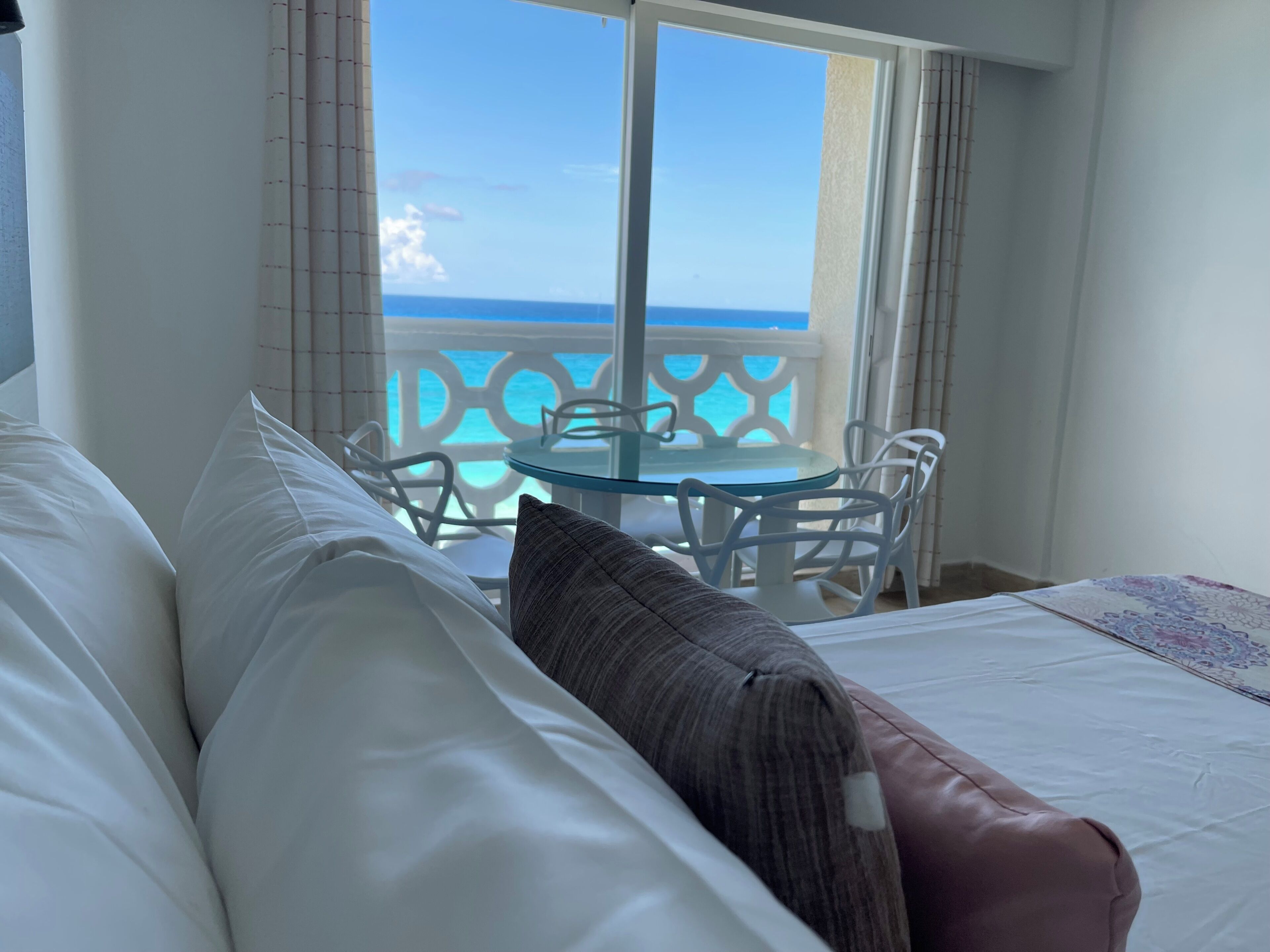 A hotel room with a bed, pillows, a glass table, chairs, and a view of the ocean through a balcony.