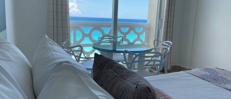 A hotel room with a bed, pillows, a glass table, chairs, and a view of the ocean through a balcony.