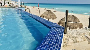 A pool adjacent to a beach with thatched umbrellas and a lifeguard on duty. - Ocean View Apartments – Increíble (Cancun)