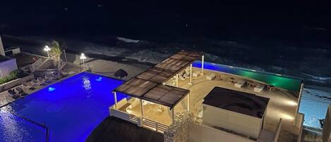 A pool area with a stone structure, a covered seating area, and a view of the sea at night.